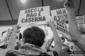 A imagem em preto e branco mostra um grupo de pessoas segurando cartazes em uma manifestação. Os cartazes, com textos em letras maiúsculas, dizem: "ESCOLA NÃO É CASERNA" e "NÃO AO PL 341/2023". As pessoas na imagem estão levantando os cartazes em um ato de protesto, defendendo a ideia de que as escolas não devem ser militarizadas. A foto é creditada ao sindicato CPERS, entidade que esteve envolvida na organização do protesto.