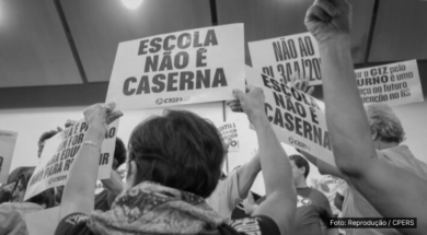 A imagem em preto e branco mostra um grupo de pessoas segurando cartazes em uma manifestação. Os cartazes, com textos em letras maiúsculas, dizem: "ESCOLA NÃO É CASERNA" e "NÃO AO PL 341/2023". As pessoas na imagem estão levantando os cartazes em um ato de protesto, defendendo a ideia de que as escolas não devem ser militarizadas. A foto é creditada ao sindicato CPERS, entidade que esteve envolvida na organização do protesto.