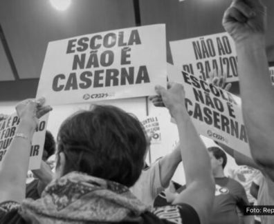 A imagem em preto e branco mostra um grupo de pessoas segurando cartazes em uma manifestação. Os cartazes, com textos em letras maiúsculas, dizem: "ESCOLA NÃO É CASERNA" e "NÃO AO PL 341/2023". As pessoas na imagem estão levantando os cartazes em um ato de protesto, defendendo a ideia de que as escolas não devem ser militarizadas. A foto é creditada ao sindicato CPERS, entidade que esteve envolvida na organização do protesto.