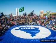 A imagem mostra um ato estudantil com centenas de pessoas reunidas em frente ao Congresso Nacional, em Brasília. No centro da imagem, há uma enorme bandeira azul com o mapa do Brasil e o logotipo da União Nacional dos Estudantes (UNE). As pessoas ao redor da bandeira estão sentadas ou agachadas, com bandeiras de movimentos estudantis. Ao fundo, vê-se a bandeira do Brasil sendo erguida.