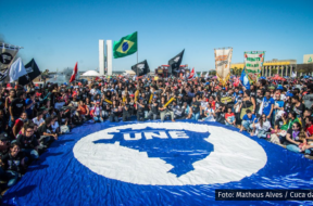 A imagem mostra um ato estudantil com centenas de pessoas reunidas em frente ao Congresso Nacional, em Brasília. No centro da imagem, há uma enorme bandeira azul com o mapa do Brasil e o logotipo da União Nacional dos Estudantes (UNE). As pessoas ao redor da bandeira estão sentadas ou agachadas, com bandeiras de movimentos estudantis. Ao fundo, vê-se a bandeira do Brasil sendo erguida.
