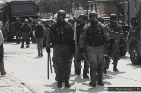 A imagem mostra uma cena de repressão policial, com vários agentes fortemente armados e equipados com escudos e cassetetes. Os policiais estão usando uniformes de proteção, incluindo capacetes e máscaras. Ao fundo, há veículos policiais, e algumas pessoas parecem estar registrando a cena com celulares. O ambiente ao redor é urbano, nas proximidades do Campus Maracanã da Universidade do Estado do Rio de Janeiro (UERJ). A legenda da foto indica que se trata de uma reprodução de uma imagem da TV Globo.