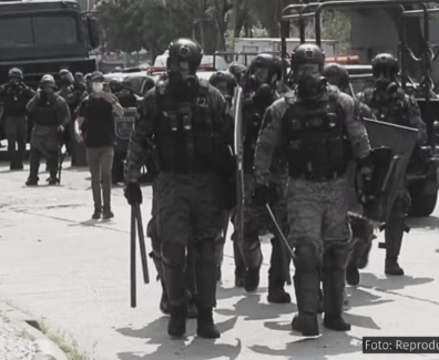 A imagem mostra uma cena de repressão policial, com vários agentes fortemente armados e equipados com escudos e cassetetes. Os policiais estão usando uniformes de proteção, incluindo capacetes e máscaras. Ao fundo, há veículos policiais, e algumas pessoas parecem estar registrando a cena com celulares. O ambiente ao redor é urbano, nas proximidades do Campus Maracanã da Universidade do Estado do Rio de Janeiro (UERJ). A legenda da foto indica que se trata de uma reprodução de uma imagem da TV Globo.