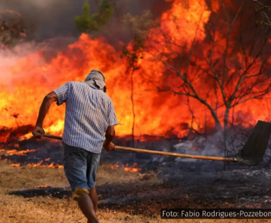 A fotografia mostra um homem de costas, usando uma camisa de manga curta e chapéu para se proteger do calor, segurando uma ferramenta enquanto tenta combater um grande incêndio florestal. As chamas tomam conta da vegetação à sua frente, em um cenário que evidencia a gravidade e o descontrole das queimadas. O fogo consome tudo ao redor, espalhando fumaça e calor intenso, com árvores secas e o solo queimado em primeiro plano. A imagem transmite a urgência e o drama da crise climática que se abate sobre o Brasil, especialmente nas regiões mais vulneráveis, como o Cerrado e a Amazônia.