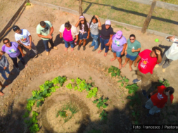 Na imagem, vemos um grupo de 14 pessoas de pé ao redor de uma horta comunitária em formato espiral, com diferentes plantas em crescimento. Eles estão em uma área cercada, ao ar livre, aparentando estarem envolvidos em um projeto de agroecologia. As pessoas estão vestidas de maneira casual e descontraída, algumas usando camisetas com símbolos da União da Juventude Comunista (UJC), da Pastoral da Juventude e de outras organizações. Todos estão de pé, em uma formação que parece abraçar a horta, com expressões de alegria, força e comprometimento. O ambiente é rural, e a horta exibe vegetação verde e saudável, representando o trabalho conjunto e o espírito de coletividade. Foto: Francesco / Pastoral da Juventude.