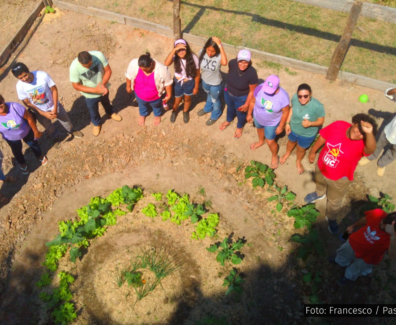 Na imagem, vemos um grupo de 14 pessoas de pé ao redor de uma horta comunitária em formato espiral, com diferentes plantas em crescimento. Eles estão em uma área cercada, ao ar livre, aparentando estarem envolvidos em um projeto de agroecologia. As pessoas estão vestidas de maneira casual e descontraída, algumas usando camisetas com símbolos da União da Juventude Comunista (UJC), da Pastoral da Juventude e de outras organizações. Todos estão de pé, em uma formação que parece abraçar a horta, com expressões de alegria, força e comprometimento. O ambiente é rural, e a horta exibe vegetação verde e saudável, representando o trabalho conjunto e o espírito de coletividade. Foto: Francesco / Pastoral da Juventude.