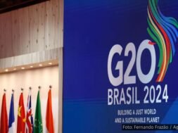 Painel azul com o logotipo do G20 Brasil 2024, acompanhado de bandeiras de países membros e a frase "Building a Just World and a Sustainable Planet".