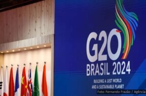 Painel azul com o logotipo do G20 Brasil 2024, acompanhado de bandeiras de países membros e a frase "Building a Just World and a Sustainable Planet".