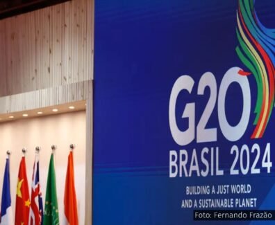 Painel azul com o logotipo do G20 Brasil 2024, acompanhado de bandeiras de países membros e a frase "Building a Just World and a Sustainable Planet".