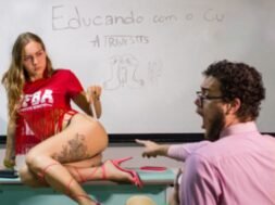 Imagem de uma performance artística, onde Tertuliana Lustosa, com camiseta vermelha, posa em cima de uma mesa de frente para um quadro branco, com um espectador expressando surpresa. No quadro, está escrito "Educando com o C* - A Travestis".