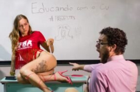 Imagem de uma performance artística, onde Tertuliana Lustosa, com camiseta vermelha, posa em cima de uma mesa de frente para um quadro branco, com um espectador expressando surpresa. No quadro, está escrito "Educando com o C* - A Travestis".