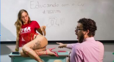 Imagem de uma performance artística, onde Tertuliana Lustosa, com camiseta vermelha, posa em cima de uma mesa de frente para um quadro branco, com um espectador expressando surpresa. No quadro, está escrito "Educando com o C* - A Travestis".