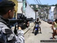 Policial aponta uma arma em direção a moradores de uma comunidade durante uma operação, cercado por pessoas. Esta imagem ilustra a violência policial que permeia o cotidiano de quem vive nas favelas. Foto: Betinho Casas Novas / Estadão.