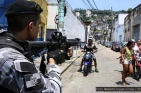 Policial aponta uma arma em direção a moradores de uma comunidade durante uma operação, cercado por pessoas. Esta imagem ilustra a violência policial que permeia o cotidiano de quem vive nas favelas. Foto: Betinho Casas Novas / Estadão.
