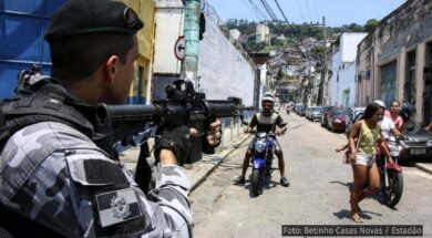 Policial aponta uma arma em direção a moradores de uma comunidade durante uma operação, cercado por pessoas. Esta imagem ilustra a violência policial que permeia o cotidiano de quem vive nas favelas. Foto: Betinho Casas Novas / Estadão.
