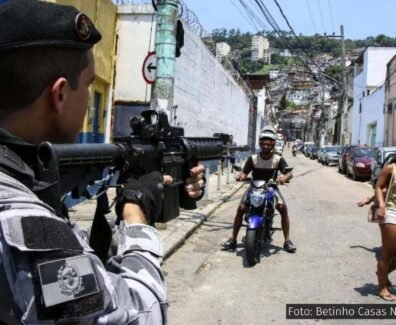Policial aponta uma arma em direção a moradores de uma comunidade durante uma operação, cercado por pessoas. Esta imagem ilustra a violência policial que permeia o cotidiano de quem vive nas favelas. Foto: Betinho Casas Novas / Estadão.