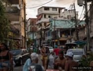 Jovens em uma rua movimentada da periferia de Salvador, com pedestres, carros e moradores sentados em mesas, ao fundo casas e postes com fiação visível. Foto: João Porfírio / Observador.