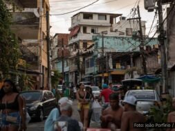 Jovens em uma rua movimentada da periferia de Salvador, com pedestres, carros e moradores sentados em mesas, ao fundo casas e postes com fiação visível. Foto: João Porfírio / Observador.