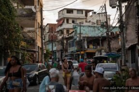 Jovens em uma rua movimentada da periferia de Salvador, com pedestres, carros e moradores sentados em mesas, ao fundo casas e postes com fiação visível. Foto: João Porfírio / Observador.
