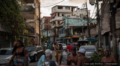 Jovens em uma rua movimentada da periferia de Salvador, com pedestres, carros e moradores sentados em mesas, ao fundo casas e postes com fiação visível. Foto: João Porfírio / Observador.