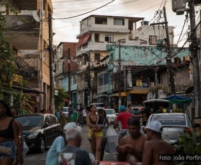 Jovens em uma rua movimentada da periferia de Salvador, com pedestres, carros e moradores sentados em mesas, ao fundo casas e postes com fiação visível. Foto: João Porfírio / Observador.