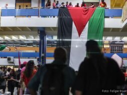 Estudantes da USP em manifestação, exibindo uma grande bandeira da Palestina pendurada em uma sacada, com grupos reunidos no andar inferior em um ato de solidariedade internacional.