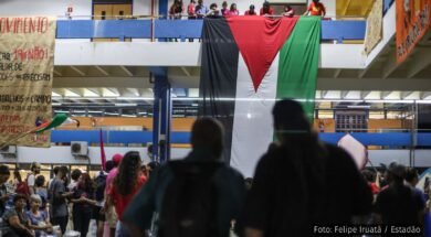 Estudantes da USP em manifestação, exibindo uma grande bandeira da Palestina pendurada em uma sacada, com grupos reunidos no andar inferior em um ato de solidariedade internacional.