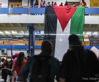 Estudantes da USP em manifestação, exibindo uma grande bandeira da Palestina pendurada em uma sacada, com grupos reunidos no andar inferior em um ato de solidariedade internacional.