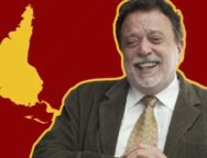 Fotografia de Theotônio dos Santos sorrindo, vestindo terno e gravata, com um mapa da América Latina invertido em relação à cartografia eurocêntrica. O mapa está em amarelo sobre um fundo vermelho listrado.