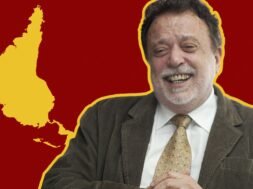 Fotografia de Theotônio dos Santos sorrindo, vestindo terno e gravata, com um mapa da América Latina invertido em relação à cartografia eurocêntrica. O mapa está em amarelo sobre um fundo vermelho listrado.