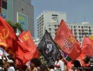 Manifestantes em ato político segurando bandeiras de movimentos sociais e organizações, como PCB e UJC, em frente a prédios urbanos. Foto: Rafa Torres / @f0tografah