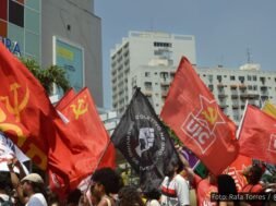 Manifestantes em ato político segurando bandeiras de movimentos sociais e organizações, como PCB e UJC, em frente a prédios urbanos. Foto: Rafa Torres / @f0tografah
