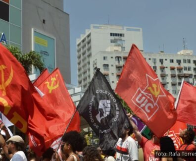 Manifestantes em ato político segurando bandeiras de movimentos sociais e organizações, como PCB e UJC, em frente a prédios urbanos. Foto: Rafa Torres / @f0tografah