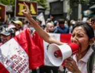 Uma mulher segura um megafone enquanto faz um discurso enérgico em um protesto no Peru. Ao fundo, outras pessoas seguram cartazes e uma bandeira peruana com inscrições exigindo justiça e liberdade.