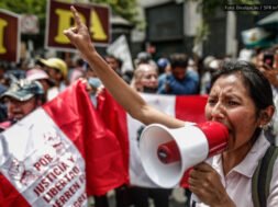 Uma mulher segura um megafone enquanto faz um discurso enérgico em um protesto no Peru. Ao fundo, outras pessoas seguram cartazes e uma bandeira peruana com inscrições exigindo justiça e liberdade.