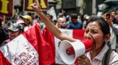 Uma mulher segura um megafone enquanto faz um discurso enérgico em um protesto no Peru. Ao fundo, outras pessoas seguram cartazes e uma bandeira peruana com inscrições exigindo justiça e liberdade.