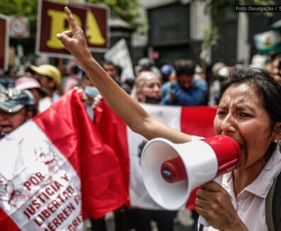 Uma mulher segura um megafone enquanto faz um discurso enérgico em um protesto no Peru. Ao fundo, outras pessoas seguram cartazes e uma bandeira peruana com inscrições exigindo justiça e liberdade.