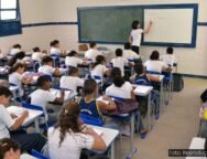 A imagem mostra uma sala de aula cheia de estudantes do ensino público, com uma professora escrevendo no quadro. O cenário representa a realidade do Novo Ensino Médio, política educacional que tem sido alvo de intensos debates sobre sua eficácia, impactos no aprendizado e desigualdades no acesso ao conhecimento.