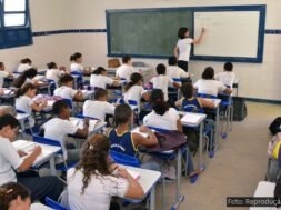 A imagem mostra uma sala de aula cheia de estudantes do ensino público, com uma professora escrevendo no quadro. O cenário representa a realidade do Novo Ensino Médio, política educacional que tem sido alvo de intensos debates sobre sua eficácia, impactos no aprendizado e desigualdades no acesso ao conhecimento.
