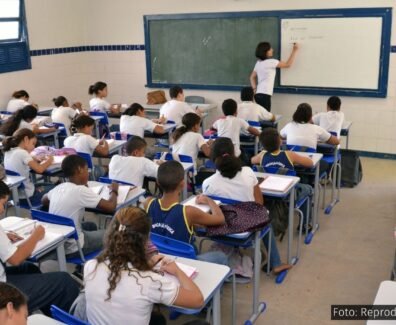 A imagem mostra uma sala de aula cheia de estudantes do ensino público, com uma professora escrevendo no quadro. O cenário representa a realidade do Novo Ensino Médio, política educacional que tem sido alvo de intensos debates sobre sua eficácia, impactos no aprendizado e desigualdades no acesso ao conhecimento.