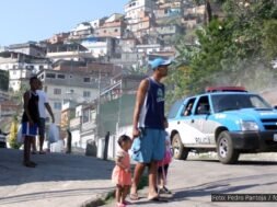 Uma cena em uma favela brasileira durante o dia, onde um homem de boné e bermuda segura a mão de uma criança pequena, enquanto outra criança caminha ao seu lado. Ao fundo, uma viatura da polícia azul e branca se aproxima, levantando poeira. Outros moradores observam a cena em meio a casas construídas em morros íngremes.