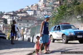 Uma cena em uma favela brasileira durante o dia, onde um homem de boné e bermuda segura a mão de uma criança pequena, enquanto outra criança caminha ao seu lado. Ao fundo, uma viatura da polícia azul e branca se aproxima, levantando poeira. Outros moradores observam a cena em meio a casas construídas em morros íngremes.