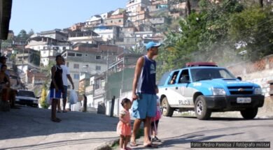 Uma cena em uma favela brasileira durante o dia, onde um homem de boné e bermuda segura a mão de uma criança pequena, enquanto outra criança caminha ao seu lado. Ao fundo, uma viatura da polícia azul e branca se aproxima, levantando poeira. Outros moradores observam a cena em meio a casas construídas em morros íngremes.