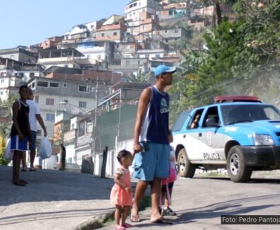 Uma cena em uma favela brasileira durante o dia, onde um homem de boné e bermuda segura a mão de uma criança pequena, enquanto outra criança caminha ao seu lado. Ao fundo, uma viatura da polícia azul e branca se aproxima, levantando poeira. Outros moradores observam a cena em meio a casas construídas em morros íngremes.