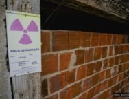 Imagem de uma entrada murada com tijolos aparentes em uma construção rústica, onde há uma placa de advertência afixada em uma viga de madeira ao lado da entrada. A placa tem o símbolo internacional de radiação (um trevo roxo sobre fundo amarelo) e contém os dizeres "RISCO DE RADIAÇÃO" com informações técnicas, incluindo localização (“Galeria G3”), data (20/09/2018), e taxa de dose medida. Acima, em verde, lê-se “ENTRADA SOMENTE DE PESSOAS AUTORIZADAS”. A estrutura aparenta abandono ou desuso e está parcialmente vedada, com sinais de deterioração, destacando o potencial perigo de contaminação radioativa no local.