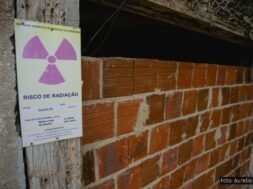 Imagem de uma entrada murada com tijolos aparentes em uma construção rústica, onde há uma placa de advertência afixada em uma viga de madeira ao lado da entrada. A placa tem o símbolo internacional de radiação (um trevo roxo sobre fundo amarelo) e contém os dizeres "RISCO DE RADIAÇÃO" com informações técnicas, incluindo localização (“Galeria G3”), data (20/09/2018), e taxa de dose medida. Acima, em verde, lê-se “ENTRADA SOMENTE DE PESSOAS AUTORIZADAS”. A estrutura aparenta abandono ou desuso e está parcialmente vedada, com sinais de deterioração, destacando o potencial perigo de contaminação radioativa no local.