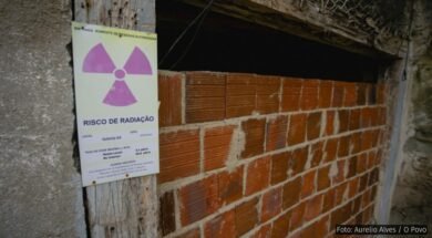 Imagem de uma entrada murada com tijolos aparentes em uma construção rústica, onde há uma placa de advertência afixada em uma viga de madeira ao lado da entrada. A placa tem o símbolo internacional de radiação (um trevo roxo sobre fundo amarelo) e contém os dizeres "RISCO DE RADIAÇÃO" com informações técnicas, incluindo localização (“Galeria G3”), data (20/09/2018), e taxa de dose medida. Acima, em verde, lê-se “ENTRADA SOMENTE DE PESSOAS AUTORIZADAS”. A estrutura aparenta abandono ou desuso e está parcialmente vedada, com sinais de deterioração, destacando o potencial perigo de contaminação radioativa no local.