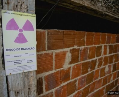 Imagem de uma entrada murada com tijolos aparentes em uma construção rústica, onde há uma placa de advertência afixada em uma viga de madeira ao lado da entrada. A placa tem o símbolo internacional de radiação (um trevo roxo sobre fundo amarelo) e contém os dizeres "RISCO DE RADIAÇÃO" com informações técnicas, incluindo localização (“Galeria G3”), data (20/09/2018), e taxa de dose medida. Acima, em verde, lê-se “ENTRADA SOMENTE DE PESSOAS AUTORIZADAS”. A estrutura aparenta abandono ou desuso e está parcialmente vedada, com sinais de deterioração, destacando o potencial perigo de contaminação radioativa no local.
