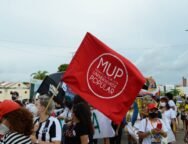 Mulher branca de cabelo loiro, usando blusa do Ceará, segura em meio a outras pessoas, uma bandeira vermelha com um círculo branco e dentro do círculo escrito MUP em letras grandes e logo abaixo Movimento por uma Universidade Popular.