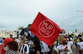 Mulher branca de cabelo loiro, usando blusa do Ceará, segura em meio a outras pessoas, uma bandeira vermelha com um círculo branco e dentro do círculo escrito MUP em letras grandes e logo abaixo Movimento por uma Universidade Popular.